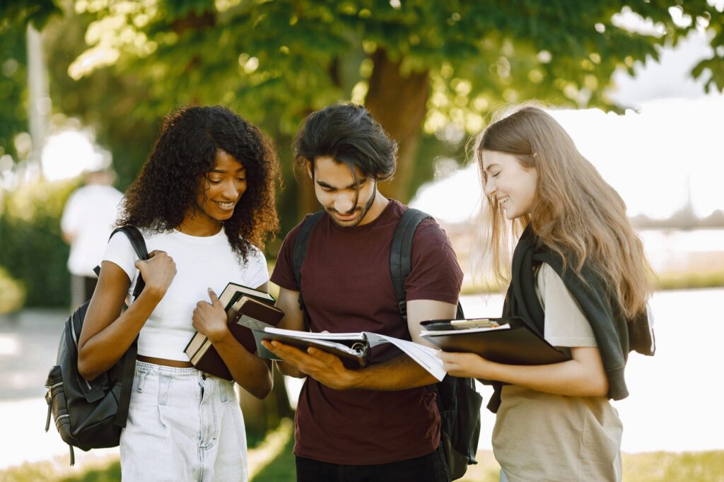 Tatlong estudyanteng internasyonal na nakatayo sa isang parke at may hawak na mga libro